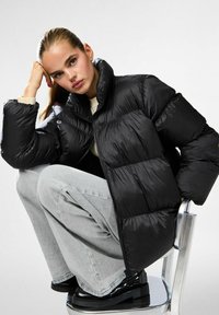 Chaqueta de plumas negra con cuello alto, diseño acolchado y cierre de cremallera. Modelo con suéter beige claro y pantalones de pierna ancha grises.