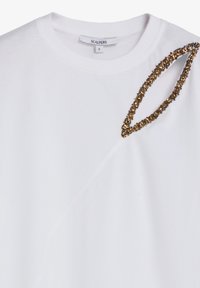 Camiseta blanca de manga corta con escote redondo, que presenta un detalle decorativo dorado a lo largo de un hombro y una suave textura de algodón.