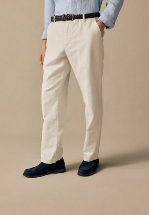 Beige Baumwollhosen mit einer lockeren Passform, kombiniert mit einem geflochtenen braunen Gürtel und dunkelblauen Wildleder-Pantoletten. Der Hintergrund ist neutral.