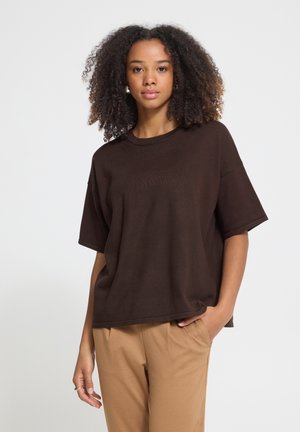 Kobieta z kręconymi włosami, ubrana w luźny ciemnobrązowy t-shirt i jasnobrązowe spodnie, stojąca z jedną ręką w kieszeni na jednolitym tle.