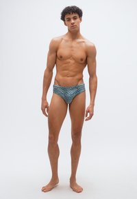 Νεαρός άνδρας στέκεται ξυπόλητος, φορώντας μπλε μαγιό με τύπωμα λεοπάρδαλης, με ουδέτερη έκφραση μπροστά σε απλό λευκό φόντο.