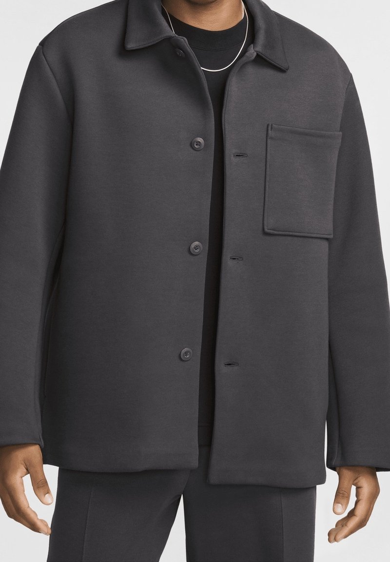 Ciemnoszara zapinana na guziki kurtka wykonana z miękkiego materiału, z kołnierzem, naszywaną kieszenią i luźnym krojem. Gładka tekstura z widocznymi przeszyciami.