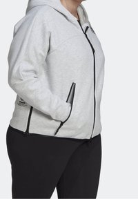 Personne portant un sweat à capuche zippé gris clair avec des détails noirs sur la fermeture éclair et un pantalon noir, debout avec une main dans la poche du sweat.