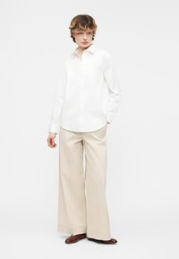 Άτομο που φοράει λευκό πουκάμισο με κουμπιά, μπεζ παντελόνι φαρδύ στο πόδι και καφέ παπούτσια, στέκεται με το ένα χέρι στην τσέπη μπροστά σε απλό φόντο.