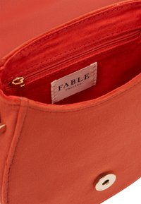 Notranjost torbice iz koralno obarvanega blaga z zadrgo in oznako "FABLE England." Ima gladko teksturo in zlatovzhodno kovinsko opremo.