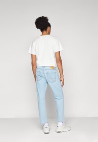 Jeans de mezclilla azul claro con un corte ajustado, que presentan bolsillos traseros cosidos y una etiqueta marrón. Combinados con una camiseta blanca sencilla y zapatillas blancas.