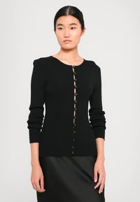 Mulher com um cardigã preto de mangas longas e canelado, ligeiramente abotoado, combinado com uma saia preta, de pé contra um fundo claro e liso.
