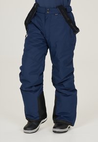 Pantalon de neige bleu marine avec des bretelles noires porté par-dessus des bottes d'hiver noires avec des semelles blanches.