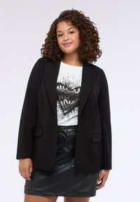 Černý blazer s upraveným střihem, s klopami a dvěma předními kapsami, nošený přes bílý grafický tričko a koženou sukni.