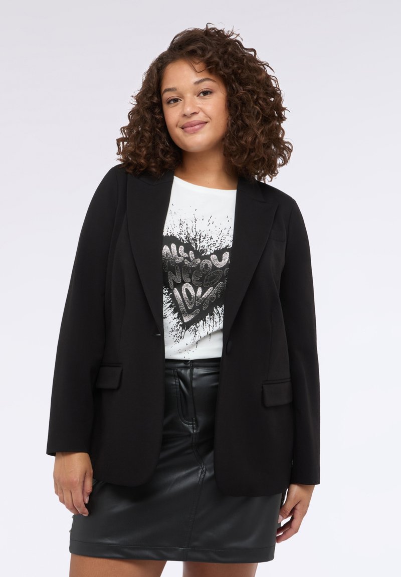 Černý blazer s upraveným střihem, s klopami a dvěma předními kapsami, nošený přes bílý grafický tričko a koženou sukni.