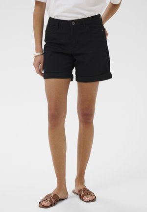 Vrouw draagt zwarte shorts met opgerolde zoom, een witte in de broek gestoken T-shirt, zilveren armband en bruine gevlochten platte sandalen, staand tegen een witte achtergrond.