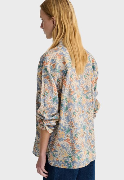 Femme aux longs cheveux blonds portant une chemise ample à manches longues à motif abstrait multicolore vue de dos.