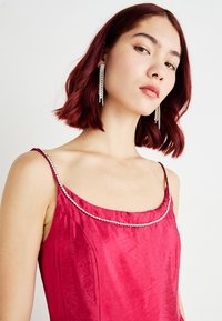 Μωβ σατέν φόρεμα με φαρδύ ντεκολτέ, διακοσμημένο με μια λαμπερή ραβδωτή ταινία από ρουμπίνια. Το μοντέλο φοράει μακριά κρυστάλλινα σκουλαρίκια σε μορφή σταγόνας.