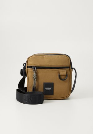 Tan canvas crossbody bag with black zipper, front pocket, and a logo patch. Features a detachable strap and a decorative zipper pull.