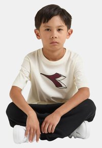 Maglietta a maniche corte color crema con grafica bordeaux, abbinata a pantaloni della tuta neri e scarpe da ginnastica bianche. Materiale in cotone morbido.