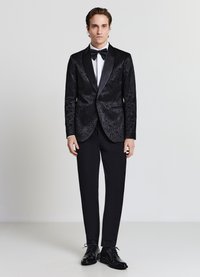 Smoking nero con tessuto strutturato, risvolti in raso, tasche frontali e accenti lucidi. Abbinato a pantaloni da cerimonia neri e scarpe formali.