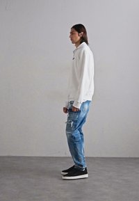 Vit sweatshirt med hög krage, kombinerad med ljusblå slitna jeans och svarta sneakers med vit sula, stående mot en grå vägg.