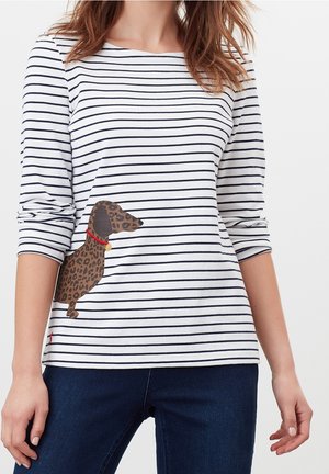 Kobieta nosząca białą koszulę z długim rękawem z cienkimi czarnymi poziomymi paskami oraz grafiką jamnika w panterkę w okolicy pasa, w połączeniu z ciemnymi jeansami.