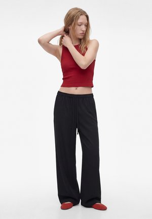 Junge Frau, die ihre Haare richtet, trägt ein rotes ärmelloses Crop Top, lockere schwarze Hosen und rote flache Schuhe, steht vor weißem Hintergrund.