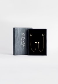 Guldtonade örhängen med cirkulära stift kopplade med delikata kedjor, visade i en svart sammetsträdd låda med en silverlogotyp på locket.