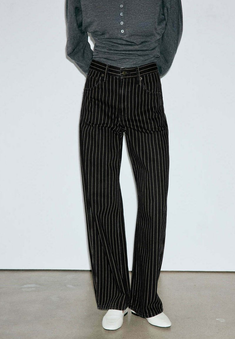 Personne portant un pantalon noir à rayures verticales blanches, un haut henley gris à manches longues et des chaussures blanches à bout carré, debout devant un fond clair.
