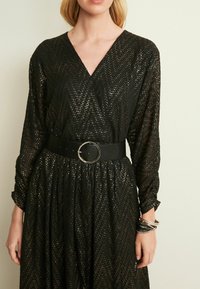 Zwarte jurk met chevronpatroon, lange mouwen, V-hals, getailleerde taille met een zwarte riem met een zilveren cirkelgesp, en een gestructureerde stof.