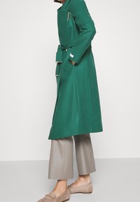 Groene wollen jas met een ritszak, een ceintuur in de taille en lange mouwen; gedragen over lichtgrijze brede broekspijpen en beige instappers.