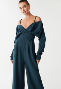 Tirkīza krāsas kombinezons gludā audumā, ar dziļu V veida izgriezumu, garām apjomīgām piedurknēm un jostu vidukļa akcentēšanai.