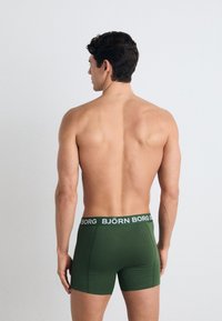 Green boxer shorts with a black waistband featuring white text, made from a smooth, stretchy material, displayed from a rear view.