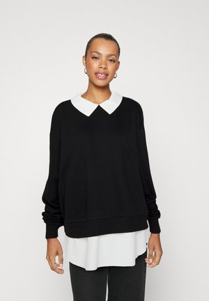 Sort sweatshirt med hvid krave, lange ærmer og afslappet pasform. Laget over en hvid top, der har en ribbet kant og struktureret stof.