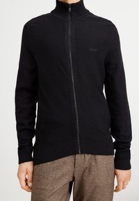 Svart zip-up-tröja med hög krage, ribbade ärmar och midja. Har subtil "BOSS"-logotyp på bröstet. Tillverkad av texturerat stickat material.