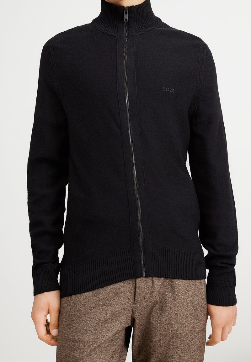 Svart zip-up-tröja med hög krage, ribbade ärmar och midja. Har subtil "BOSS"-logotyp på bröstet. Tillverkad av texturerat stickat material.