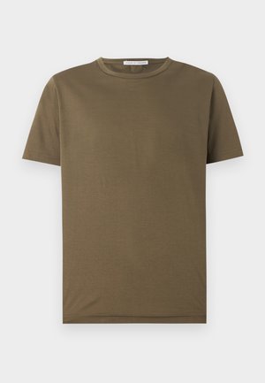Homme aux cheveux bouclés portant un t-shirt vert olive, un pantalon beige à jambes larges avec une ceinture noire, des baskets blanches, debout les mains dans les poches.