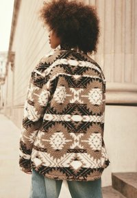 Fleecejacka med ett geometriskt mönster i brunt, beige och vitt. Designen inkluderar tribal-inspirerade motiv och en mysig textur.