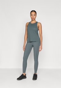 Sötétzöld sportos tank top és leggings rugalmas anyagból, laza szabással és lapos varratokkal tervezve, fekete sportcipővel párosítva.