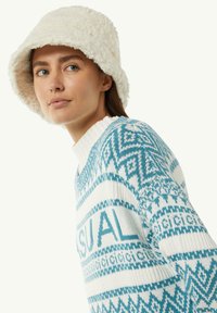 Biały, puszysty kapelusz w formie wiadra o miękkiej fakturze. Niebieski i biały sweter w wzory geometryczne z napisem "CASUAL" na klatce piersiowej.