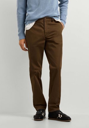 Man draagt bruine rechte broek, zwart-witte sneakers en een blauwe gebreide trui met een wit overhemd eronder.