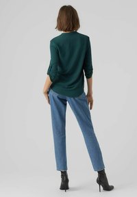 Tee in Teal mit langen Ärmeln und gerippter Textur, kombiniert mit hellblauen Jeans und schwarzen, knöchelhohen Stiefeln mit Absatz. Ansicht von hinten.