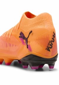 Oranssi jalkapallokengän päällinen, jossa on tekstuuri, vaaleanpunaiset liukuvärjätyt yksityiskohdat, musta ulkopohja ja rohkea Puma-logo, jossa teksti "Play Free Future".