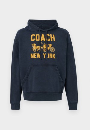 Tumszils kapuces sporta krekls ar priekšējo kabatu, dzeltenu tekstu "COACH NEW YORK" un zirgu pajūga attēlu uz krūtīm.