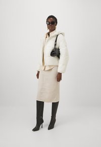 Giacca bianca piumosa con cappuccio, camicia beige, gonna beige fino al ginocchio e stivali neri alti fino alla coscia con punta; accessorio: piccola borsa nera.