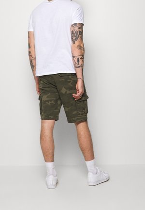 Mand stående med ryggen mod kameraet, iført hvid t-shirt, camouflage cargo shorts, hvide sokker, hvide sneakers og synlige arm-tatoveringer.