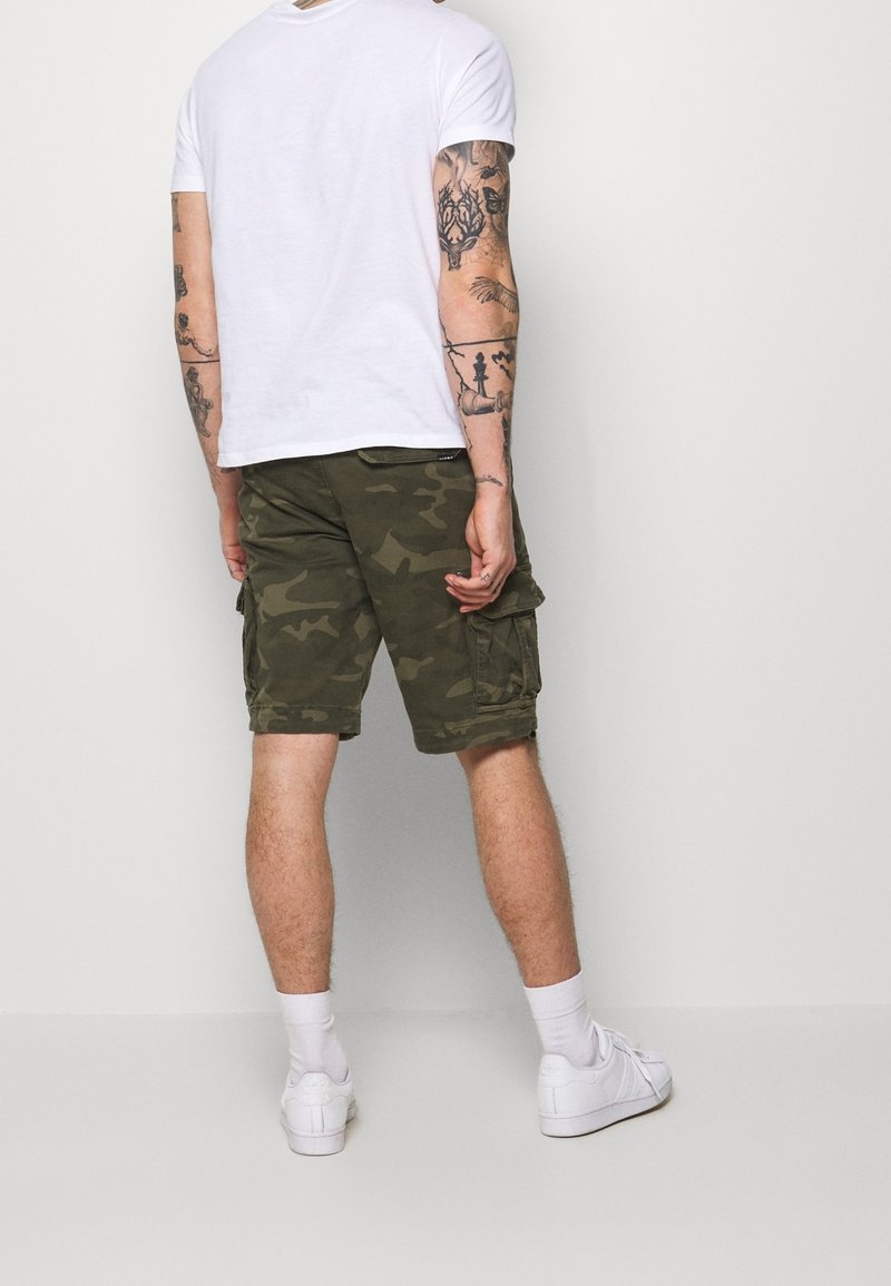 Homme debout, dos à la caméra, portant un t-shirt blanc, un short cargo camouflage, des chaussettes blanches, des baskets blanches et des tatouages visibles sur les bras.