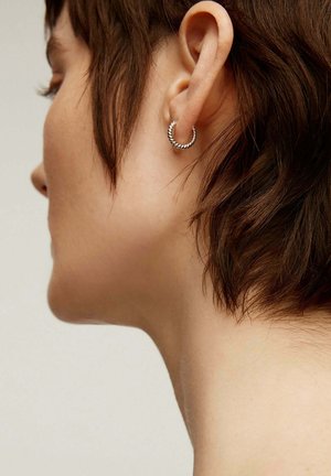 Pendiente de aro de plata con un diseño retorcido, montado en la oreja. Un cabello corto enmarca la cara, resaltando el detalle del pendiente.