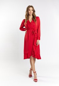Rochie roșie cu volane, mâneci lungi și talie legată. Textură fină, cu un tiv până la genunchi, completată de tocuri roșii asortate.
