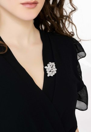 Black fabric top with a V-neckline, featuring a silver floral brooch made of textured leaves accenting the chest area. Sheer details on sleeves.