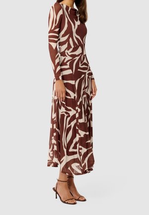 Kobieta stojąca bokiem, ubrana w midi sukienkę z długim rękawem o abstrakcyjnym brązowo-beżowym wzorze oraz szpilki z paskiem wokół kostki.