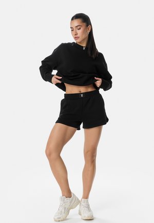 Žena nosící černou krátkou mikinu s dlouhým rukávem a černé šortky s bílými teniskami, lehce zvedající mikinu, aby ukázala tetování na břiše.