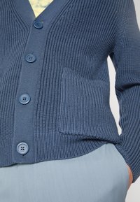 Blå stickad kofta med texturerad ribbning, som har stora blå knappar, en framficka på vänster sida och en v-ringning.