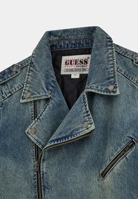 Close-up van een lichtblauwe spijkerjacket met zichtbare kraag, rits en knopen, met een "GUESS Originals" label aan de binnenkant.